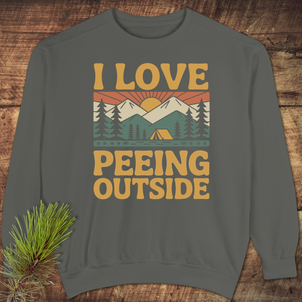 I Love Peeing Outside Sweatshirt featuring a retro outdoor scene with a sunrise, snow-capped mountains, and pine trees. Includes a bold mustard-yellow slogan, showcased on a wooden surface with pine sprig.