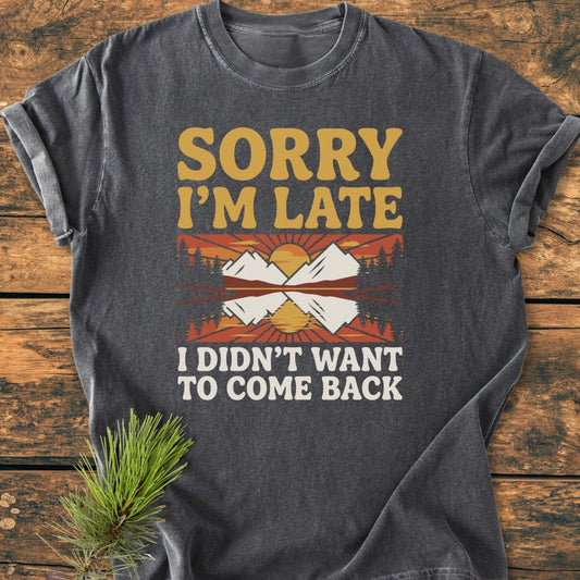 Sorry I'm Late - Vintage Wash Tee features a retro sun and mountain graphic on a charcoal-gray, short-sleeve t-shirt, laid flat with a pine sprig, emphasizing comfort and durability.