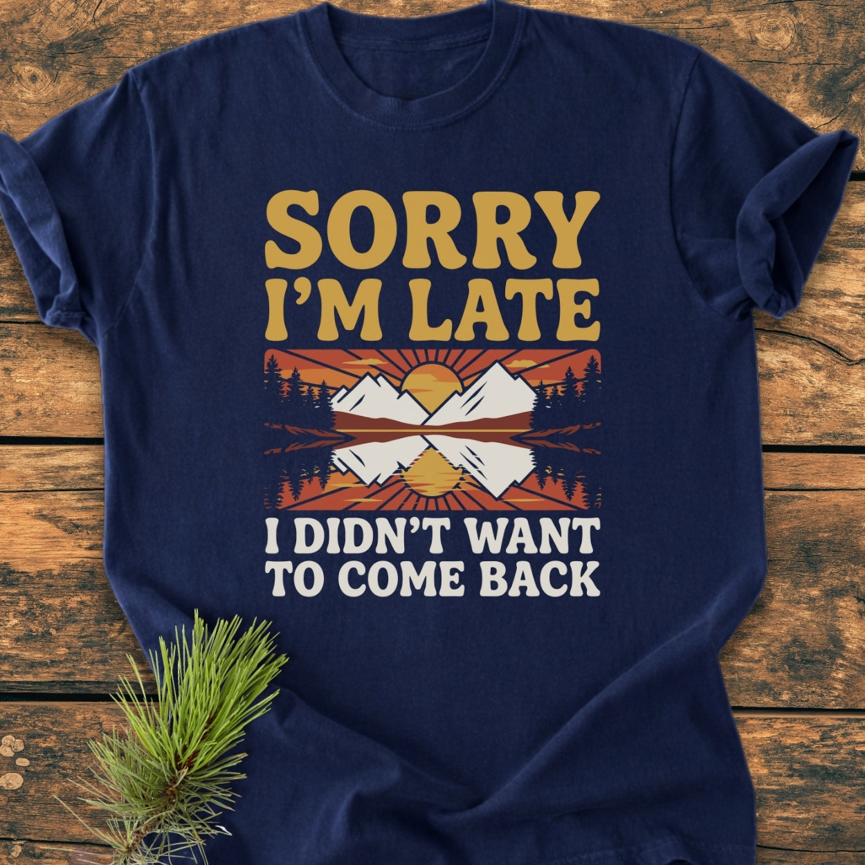 Sorry I'm Late - Vintage Wash Tee featuring a retro sun and mountains graphic with humorous text, laid flat on wood with a pine sprig, showcasing its relaxed fit and garment-dyed fabric.