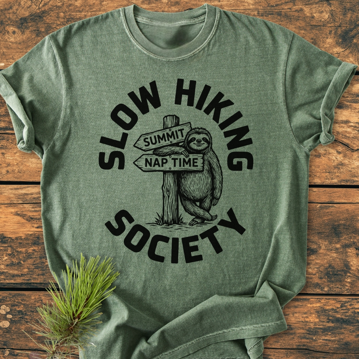 Slow Hiking Society - Vintage Wash Tee features a cartoon sloth on a trail sign, laid on a wooden surface with a pine branch, highlighting its relaxed fit and soft fabric.