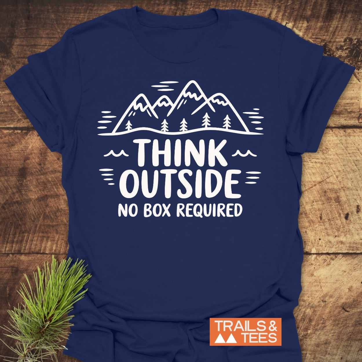 Navy-blue Think Outside T-Shirt featuring a white graphic of mountains and trees, laid flat on wood. Includes TRAILS & TEES logo, showcasing adventure-ready design and sustainable craftsmanship.