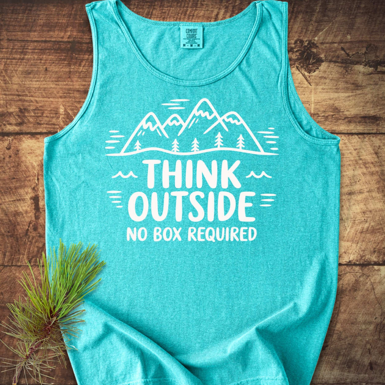 Think Outside Tank Top featuring a mountain-and-trees graphic and slogan. Laid on wood with pine needles, showcasing its relaxed fit and eco-friendly cotton material for sustainable fashion enthusiasts.