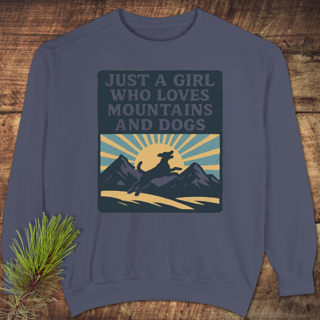 Slate-blue Just A Girl Who Loves Dogs Sweatshirt featuring a mountain and dog silhouette graphic, laid on a wooden surface. Made from soft, garment-dyed cotton-poly blend for luxurious comfort.