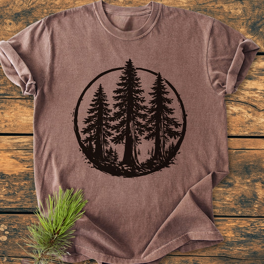 Redwood Trees - Vintage Wash Tee features a stylized forest scene on a brown short-sleeved T-shirt, laid flat on a wooden surface with a pine sprig beside it, highlighting its rustic charm.