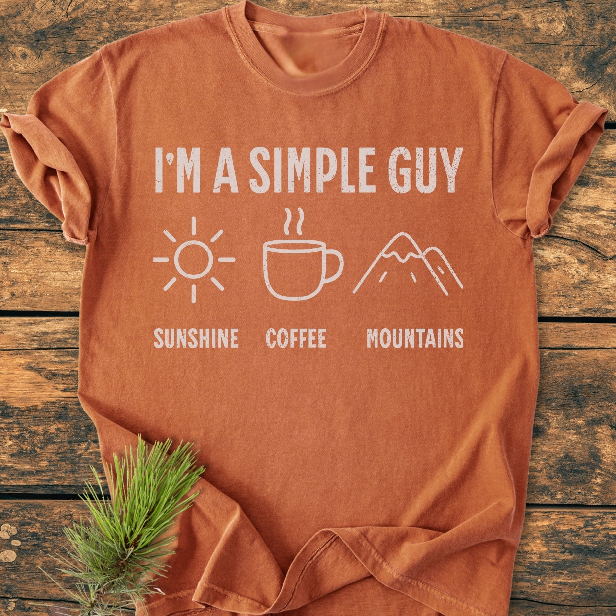 I'm A Simple Guy - Sunshine Coffee Mountains - Vintage Wash Tee displayed on wooden plank, featuring white distressed lettering with icons of sun, coffee mug, and mountains, highlighting its relaxed, soft-washed fit.