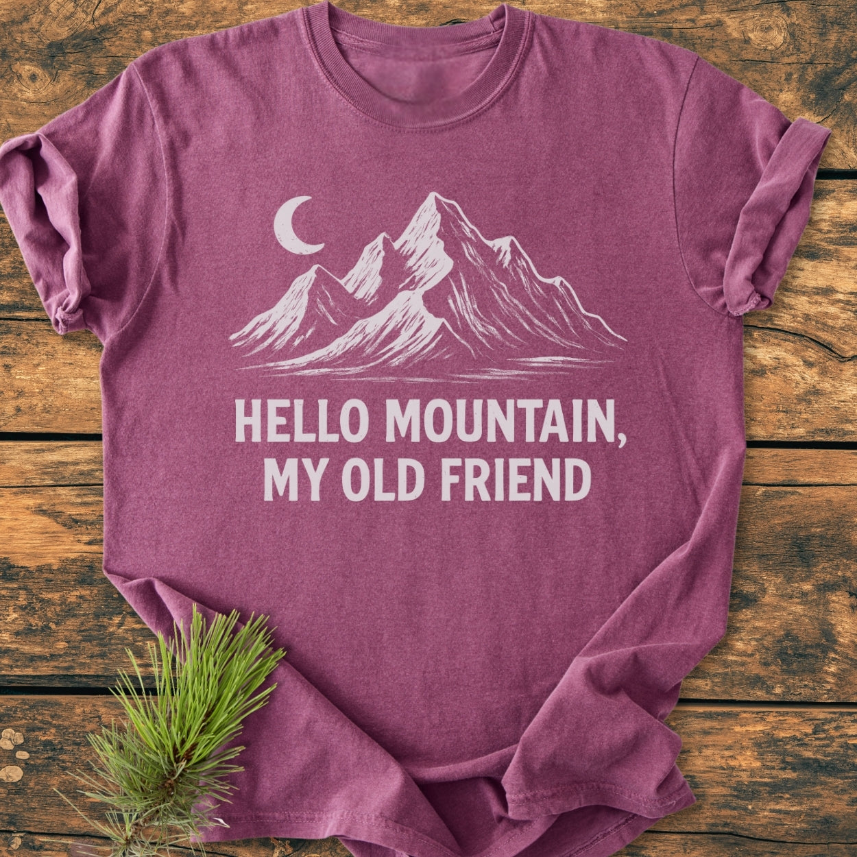 Hello Mountain My Old Friend - Vintage Wash Tee featuring a white mountain silhouette and crescent moon design, laid on rustic wood with a pine sprig, showcasing its relaxed, cozy fit.