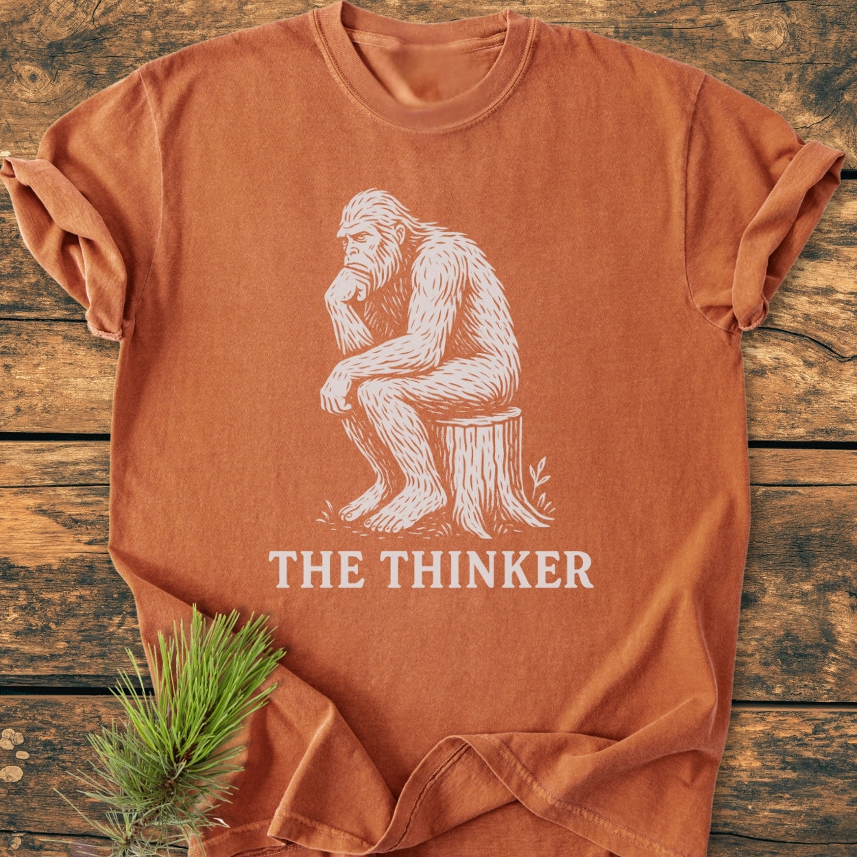Bigfoot The Thinker - Vintage Wash Tee features a rust-orange hue with a white sketch of Bigfoot in a thinker pose. Laid on wood, it’s a soft, relaxed-fit garment-dyed t-shirt.