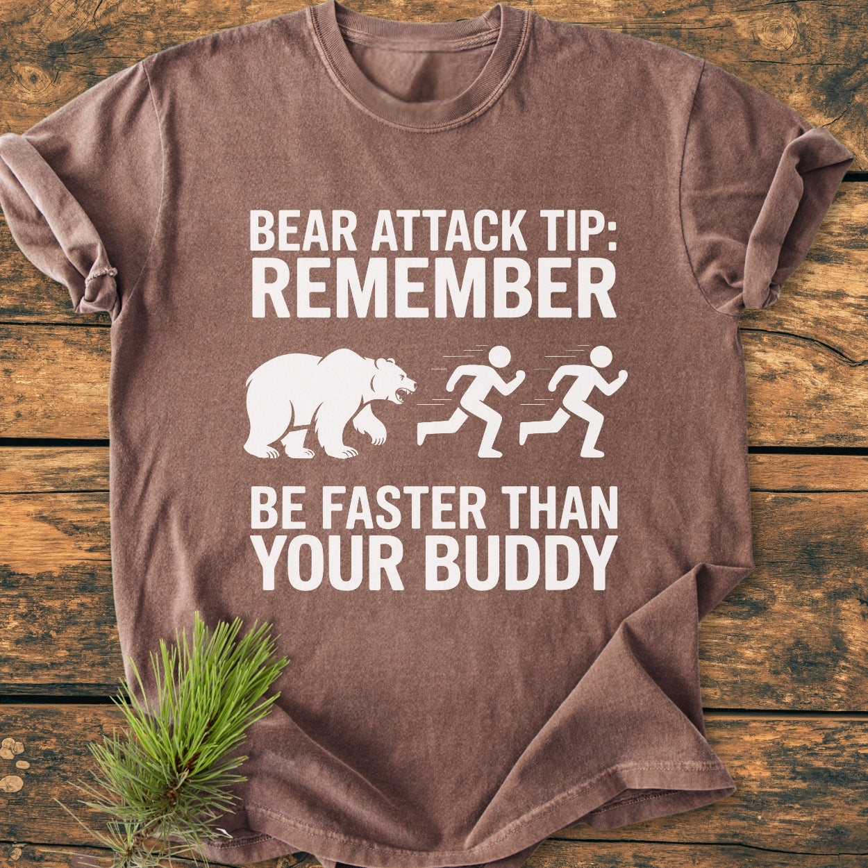 Bear Attack Tip Be Fast - Vintage Wash Tee featuring a humorous bear graphic and text, laid on a wooden surface with a pine sprig, highlighting its relaxed fit and soft texture.