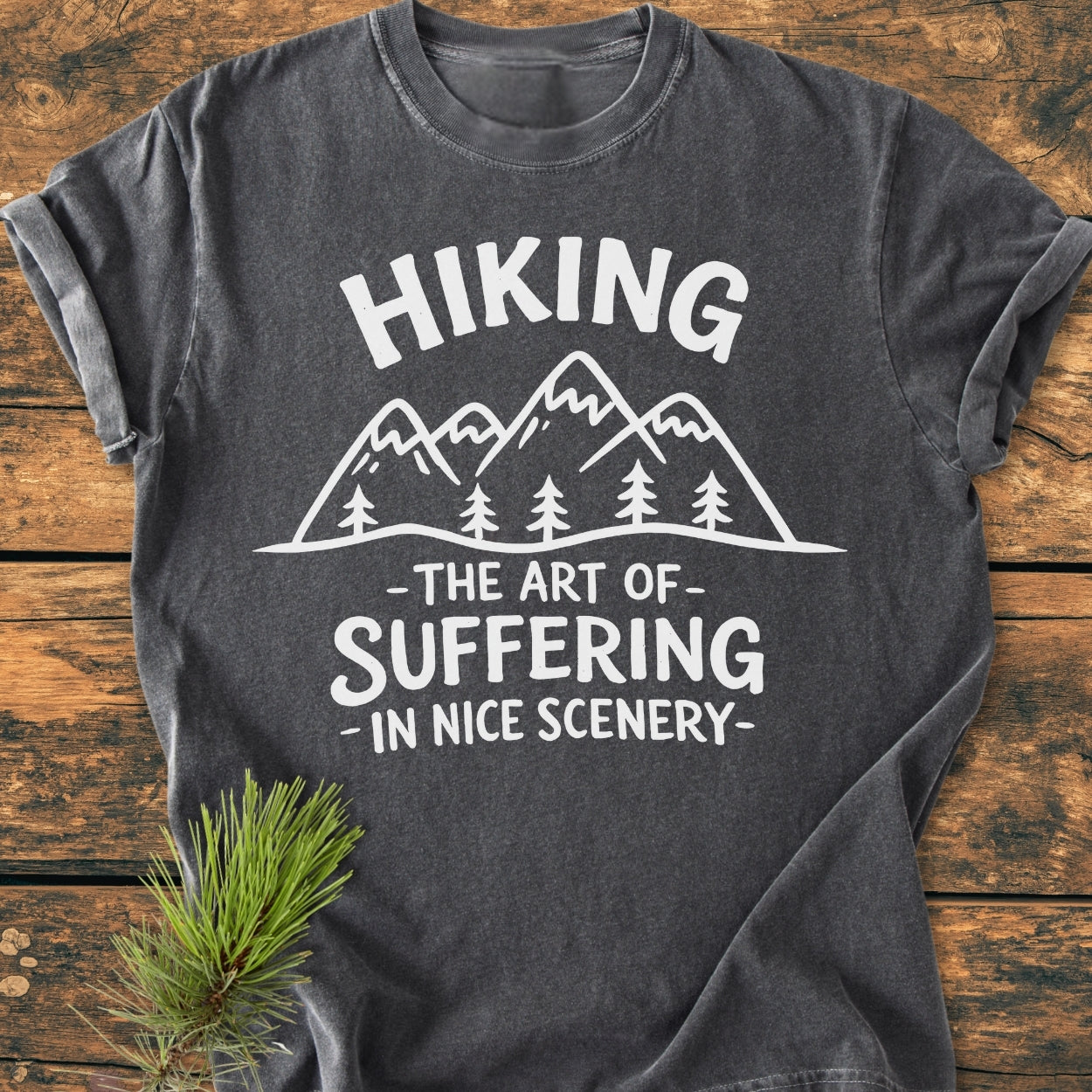 Dark-gray Hiking. The Art Of Suffering In Nice Scenery - Vintage Wash Tee featuring white stylized mountains and trees graphic on soft, durable 100% ring-spun cotton, laid on wood surface with pine sprig.