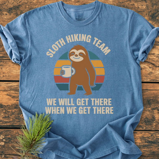 Sloth Hiking Team - Vintage Wash Tee features a cartoon sloth with a mug against a stylized sunset, laid on wood with a pine branch. Cozy, durable, relaxed fit in heather-blue.