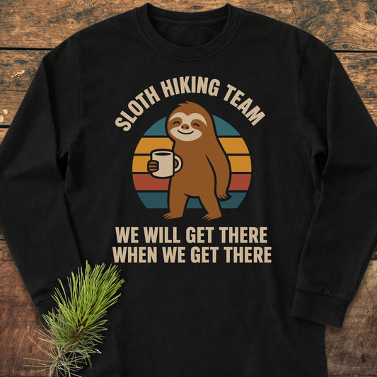 Sloth Hiking Team Long-Sleeve Tee features a cartoon sloth with a coffee mug, retro sunset, and the slogan. Laid on wood with a pine sprig, showcasing its casual, relaxed fit.