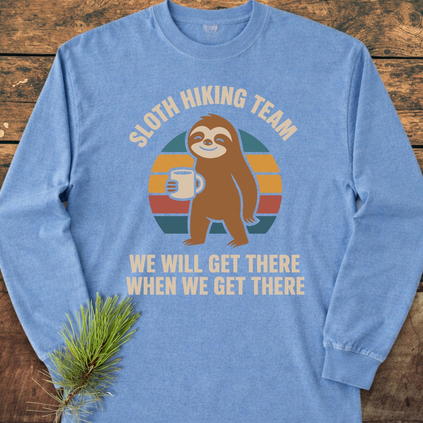 Sloth Hiking Team Long-Sleeve Tee featuring a retro-style smiling sloth with a coffee mug on a sunset-striped background, laid flat on a wooden surface with a pine sprig nearby.
