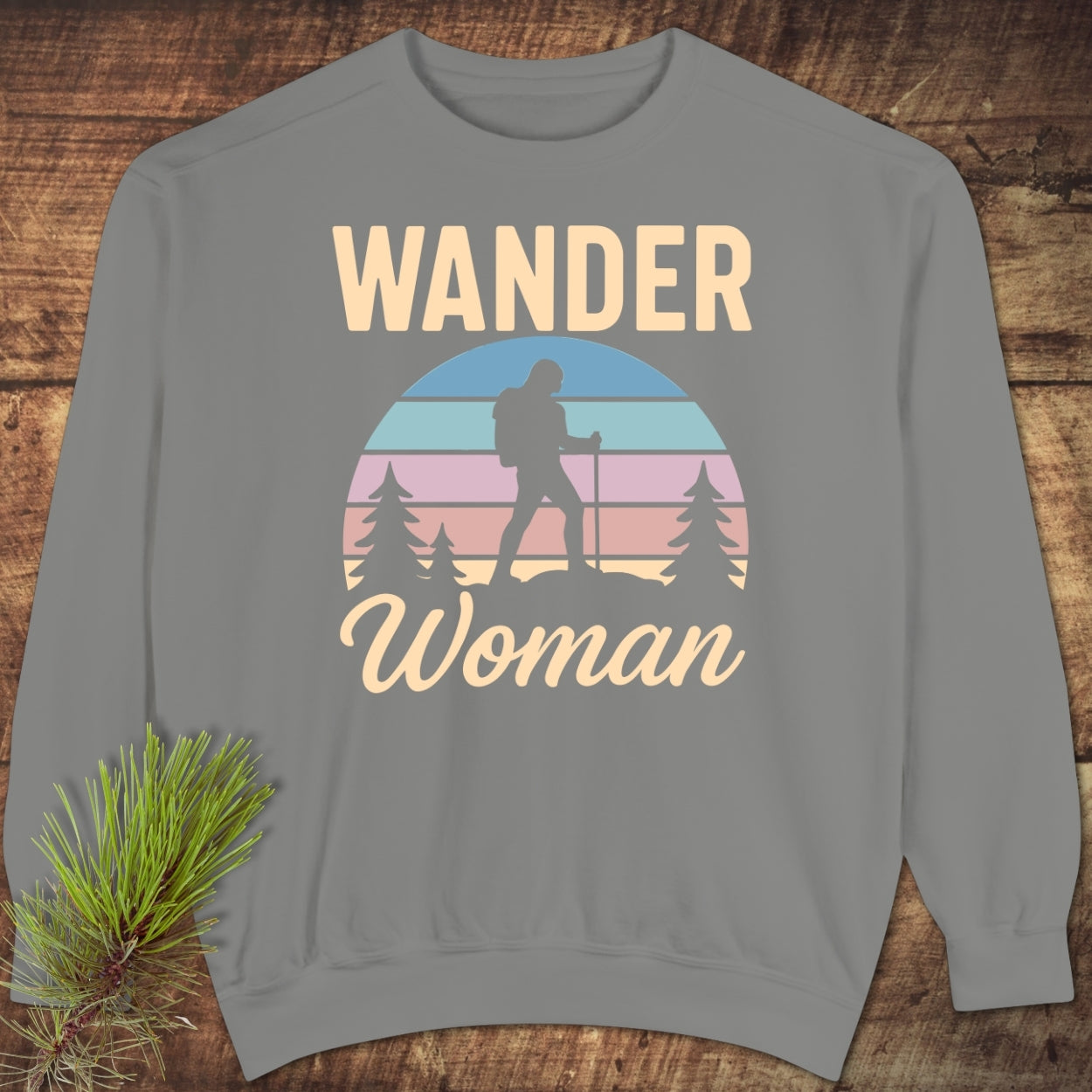 Wander Woman Retro Sweatshirt featuring a gray crewneck design with WANDER and Woman text, sunset-style illustration of a hiker amidst pine trees, laid on rustic wood.