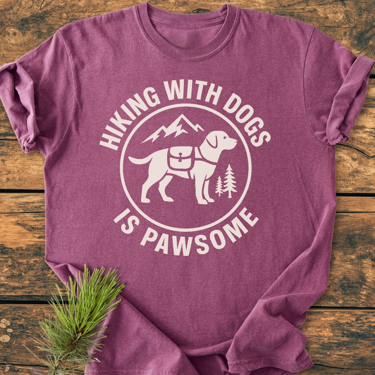Hiking With Dogs Is Pawsome - Vintage Wash Tee features a white circular graphic with a dog, mountains, and trees, laid on rustic wood. Ideal for cozy, relaxed everyday wear.