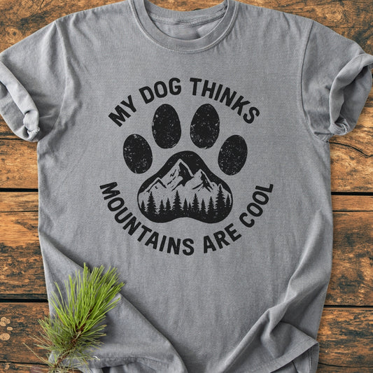 Dog Thinks Mountains Are Cool - Vintage Wash Tee featuring a large dog paw with a mountain range and pine trees, printed in black on a heather-gray T-shirt, laid flat on wood.