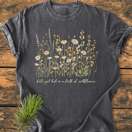 Let's Get Lost In A Field Of Wildflowers - Vintage Wash Tee features a line-drawing bouquet of daisies and blossoms with cursive text, laid on a wooden surface, embodying comfort and personalized style.