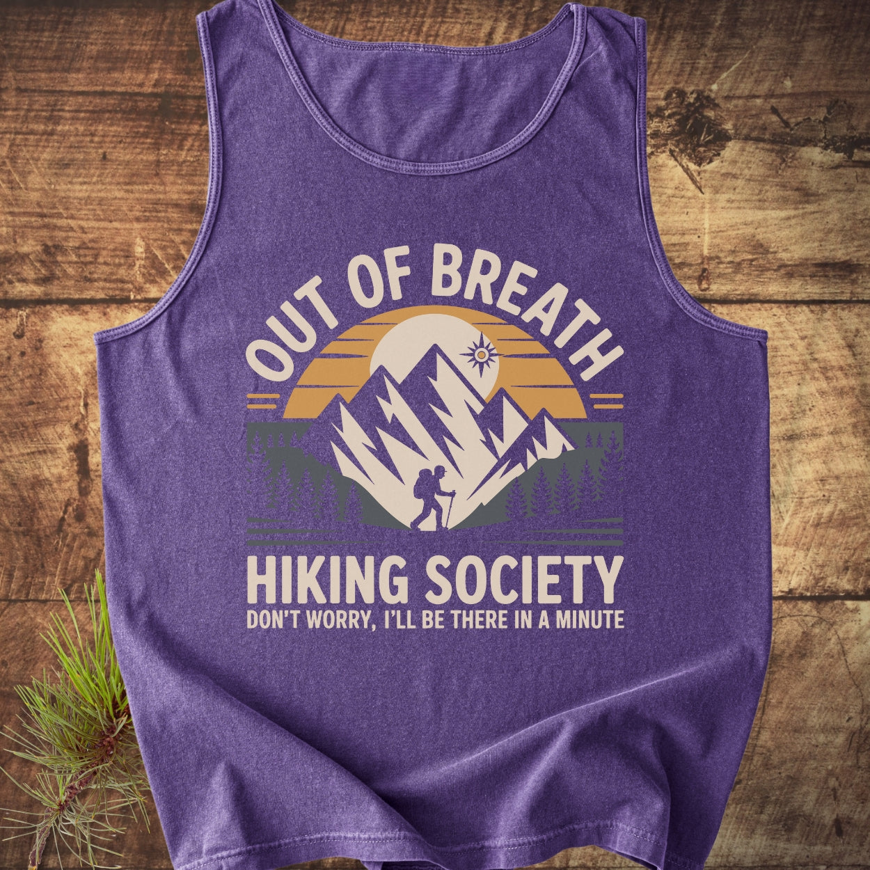 Out Of Breath Hiking Society Tank Top featuring a hiker silhouette with mountain and sun graphic, laid on wood. Accompanied by a pine branch, highlighting the outdoor adventure theme.