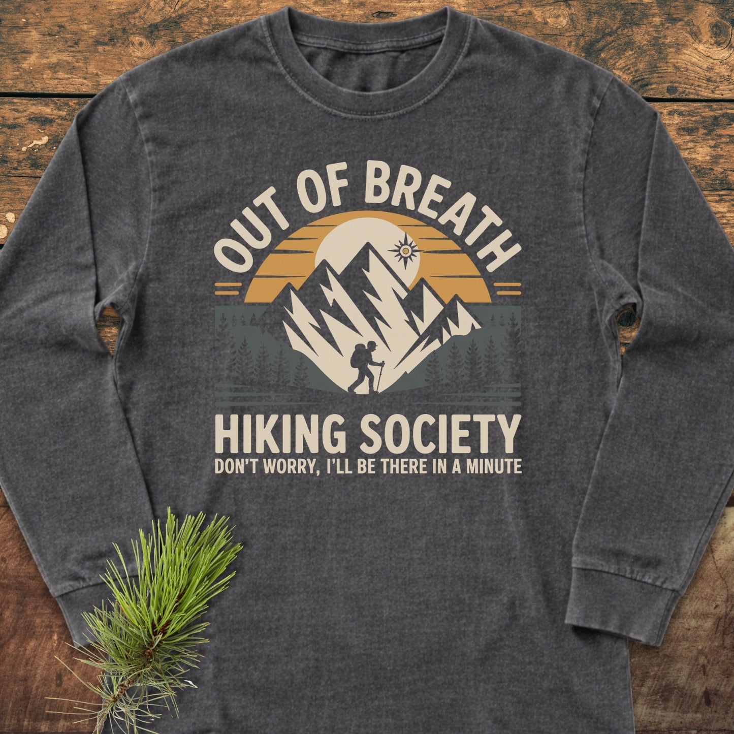 Out Of Breath Hiking Society Long-Sleeve Tee featuring a stylized mountain scene with a hiker silhouette, laid on wood with a pine sprig; made from soft, 100% ring-spun cotton.
