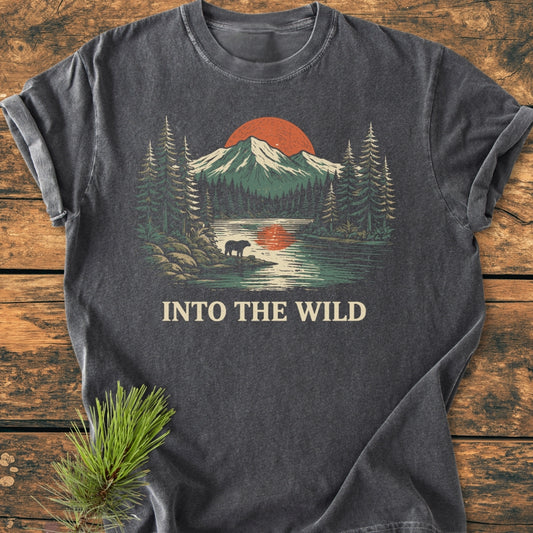 Into The Wild - Vintage Wash Tee featuring a stylized wilderness scene with mountains, a red sun, pine trees, a bear by a lake, and the caption. Laid on a wooden surface.