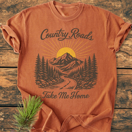 Country Roads - Vintage Wash Tee folded on wood surface, featuring a rustic road graphic with pine trees, mountains, and sun. Text reads “Country Roads, Take Me Home.” Beside the tee is an evergreen sprig.