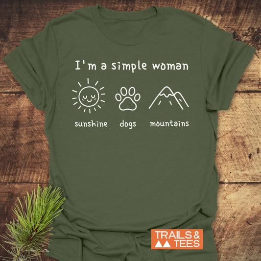 I'm A Simple Woman - Sunshine, Dogs, Mountains T-Shirt on rustic wood, featuring white icons of a sun, paw print, and mountains. Trails & Tees logo in corner, exuding outdoor adventure spirit.