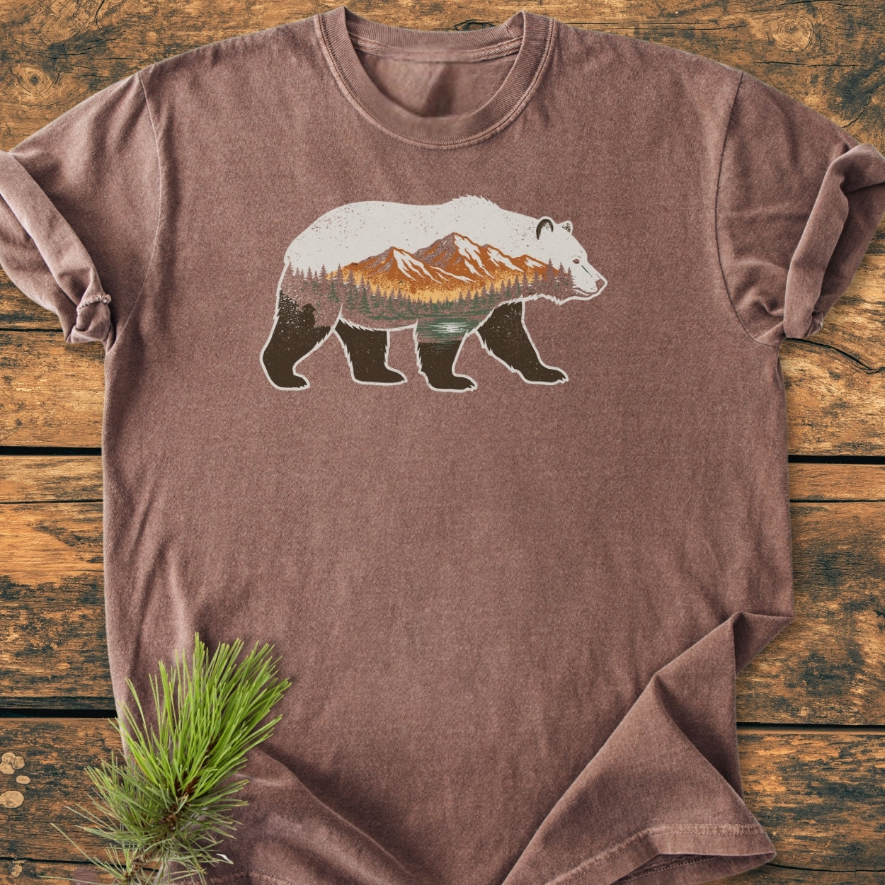 Mountain Within - Vintage Wash Tee: A brown, short-sleeve T-shirt with a bear silhouette showcasing a mountain landscape, laid flat on a wooden surface with a pine sprig nearby. Made from soft, 100% ring-spun cotton.