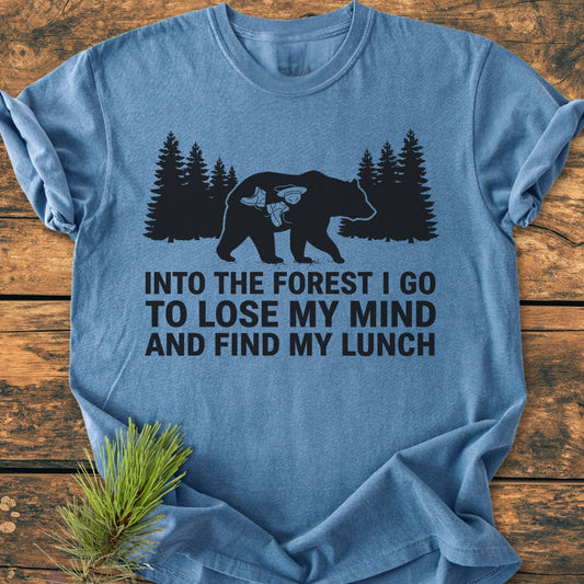 Into The Forest Bear - Vintage Wash Tee features a black bear graphic with hiking gear icons, overlaid with a nature-inspired slogan, displayed on a light-blue short-sleeve T-shirt on a wooden surface.