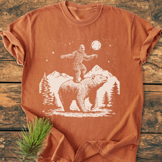 Bigfoot And Bear - Vintage Wash Tee features a wilderness scene with a bear and Sasquatch under a moon, printed on a rust-orange, relaxed fit, garment-dyed, 100% ring-spun cotton T-shirt.