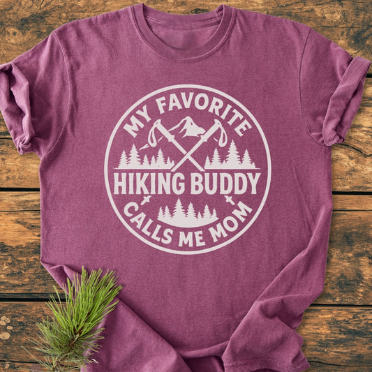 Hiking Buddy Mom - Vintage Wash Tee features a white circular graphic with crossed hiking poles, mountain, and tree line on a maroon short-sleeve T-shirt, laid on a wooden surface with a pine sprig.