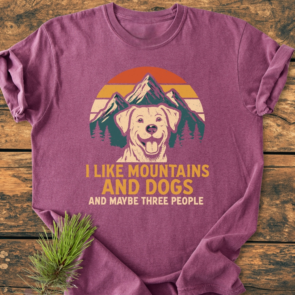 I Like Mountains And Dogs - Vintage Wash Tee features a mountain, sunset, and dog print with a playful slogan on a purple-mauve shirt, laid on wood, with a pine sprig beside it.