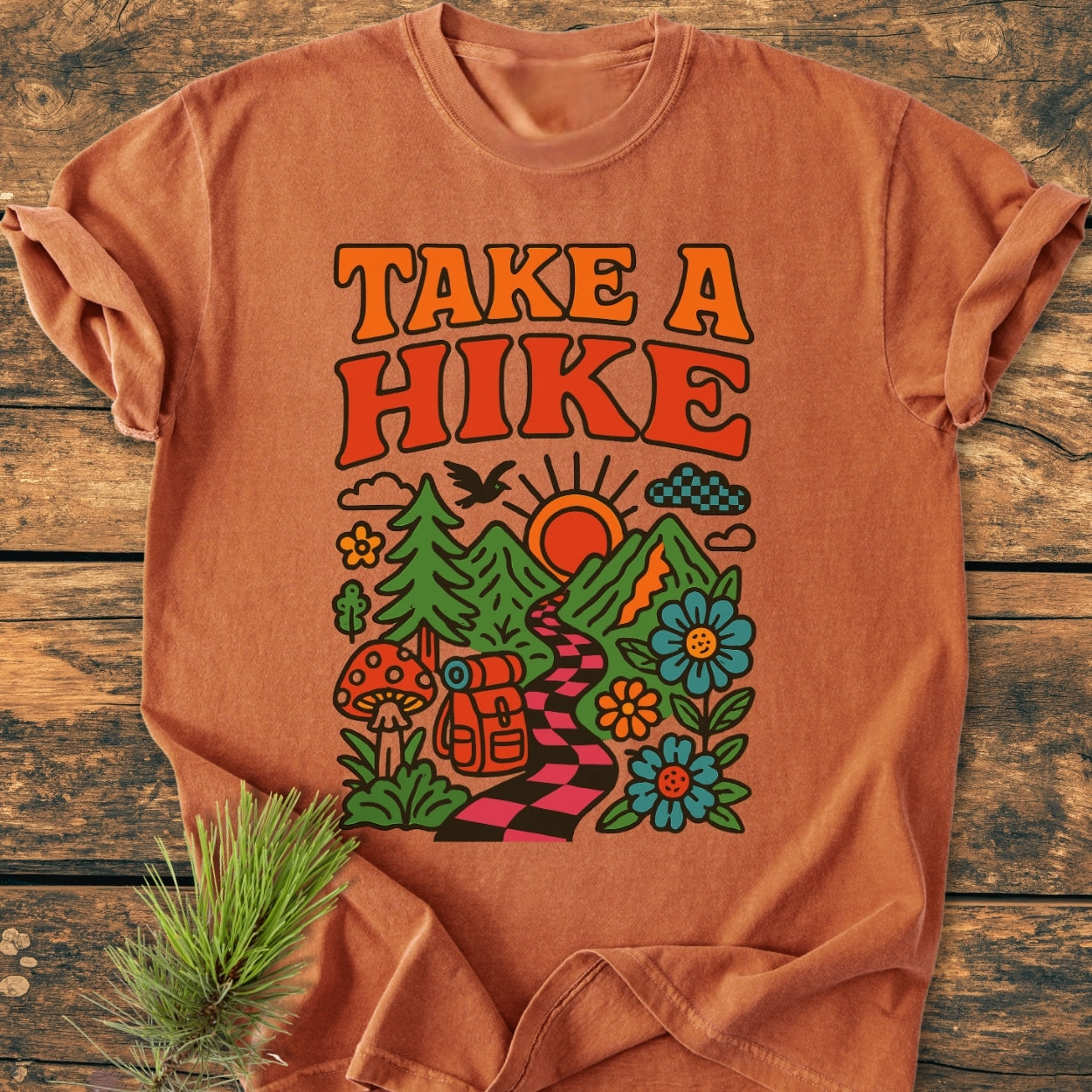 Take A Hike Funky - Vintage Wash Tee featuring a retro-style graphic with nature scene details, laid on wood. Includes checkerboard path, mountains, sun, trees, flowers, and a hiker's backpack.