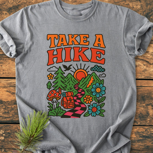 Take A Hike Funky - Vintage Wash Tee featuring retro TAKE A HIKE slogan and a colorful nature scene with mountains, trail, and backpack. Laid flat on a wooden surface with pine sprig.