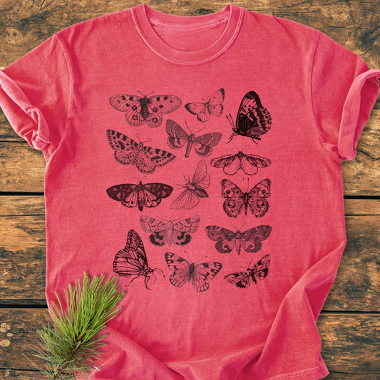 Vintage Butterfly Tattoo - Vintage Wash Tee featuring black line-art illustrations of moths and butterflies on a heathered pink/red shirt, laid on a rustic wooden surface with a sprig of pine needles.
