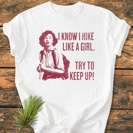 Hike Like A Girl - Vintage Wash Tee featuring a red vintage-style illustration of a woman hiking with bold text. Laid on a wooden surface with a pine sprig accent.