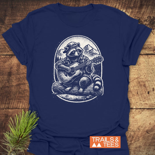Guitar Playing Raccoon T-Shirt featuring a raccoon in a backward cap and flannel, playing guitar on a log against a mountain backdrop, embodies outdoor adventure. Includes TRAILS & TEES logo.