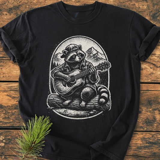 Guitar Playing Raccoon - Vintage Wash Tee features a raccoon illustration with a cap and plaid shirt playing guitar, set against a mountain scene. Laid flat on wood, showcasing its relaxed fit.