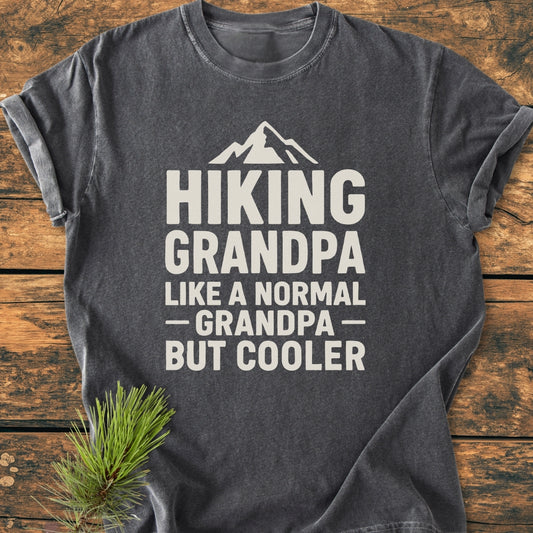 Hiking Grandpa - Vintage Wash Tee features a stylized mountain peak graphic with a catchy slogan on a dark-gray, short-sleeve shirt, laid on wood with a pine sprig nearby.