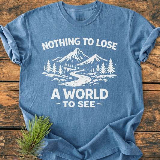 Nothing To Lose - Vintage Wash Tee with a mountain-and-forest graphic and slogan, laid flat on wood with a pine sprig beside it, showcasing soft-blue, relaxed fit, and durable stitching.