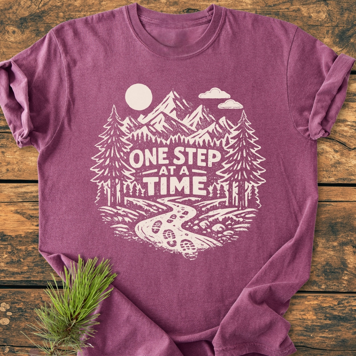 One Step At A Time - Vintage Wash Tee features a heather-purple design with a scenic mountain print and motivational slogan, laid flat on wood, showcasing its cozy, garment-dyed fabric and relaxed fit.