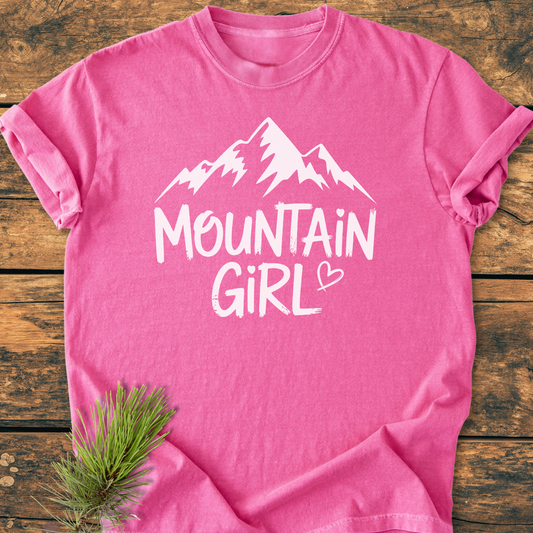 Mountain Girl - Vintage Wash Tee: A pink cotton T-shirt with a white mountain silhouette and “MOUNTAIN GIRL” text, laid flat on wood with pine needles for a rustic touch.