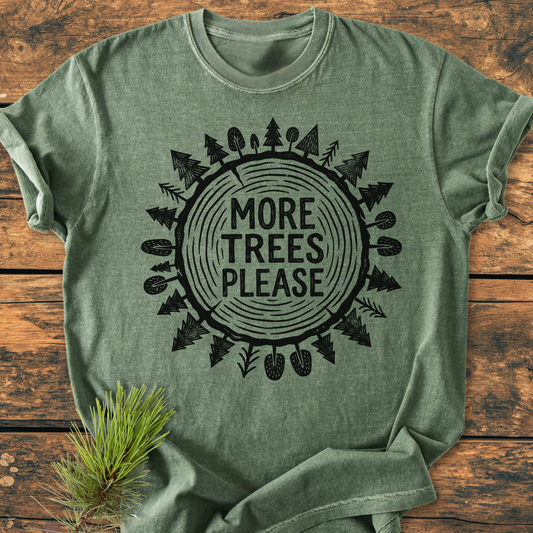More Trees Please - Vintage Wash Tee: Green heather-style tee with a tree trunk design and “MORE TREES PLEASE” text, laid on wood. A pine sprig accents the soft, relaxed fit shirt.
