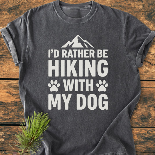 Charcoal-gray I'd Rather Be Hiking With My Dog - Vintage Wash Tee displayed on wood with pine sprig, featuring white print, mountain silhouette, and paw prints, highlighting its cozy, relaxed fit and durable stitching.