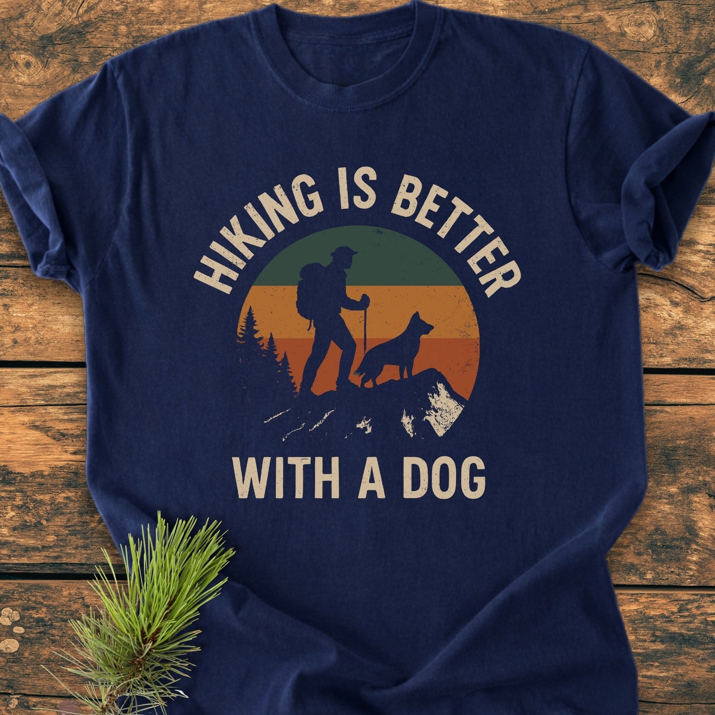 Hiking Is Better With A Dog - Vintage Wash Tee features a sunset graphic of a hiker and dog. Laid on wood with a pine sprig, highlighting its soft, durable, and relaxed fit.