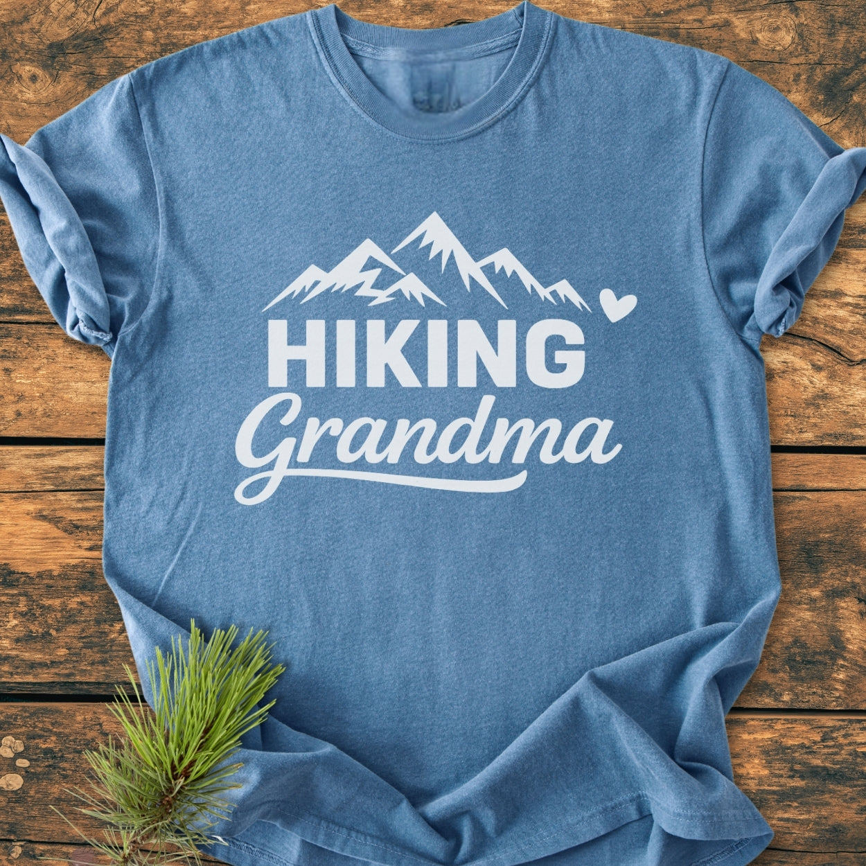 Hiking Grandma - Vintage Wash Tee: Blue crew-neck T-shirt with a mountain range and “HIKING Grandma” print, laid flat on wood, accented by a pine sprig for an outdoorsy touch.