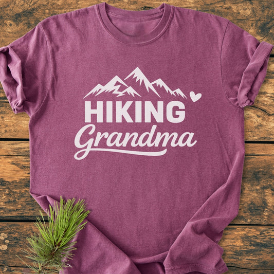 Hiking Grandma - Vintage Wash Tee displayed on wood, featuring a white mountain graphic and text. Includes a heart detail. Pine branch accents the bottom, highlighting the garment's relaxed, soft-washed texture.