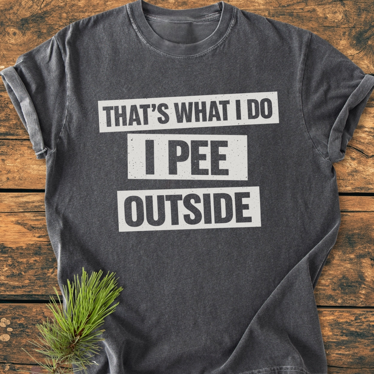 That's What I Do. I Pee Outside - Vintage Wash Tee displayed on wood, featuring a bold white slogan. A pine sprig suggests an outdoor theme, highlighting its relaxed, durable fit and soft-washed, garment-dyed fabric.