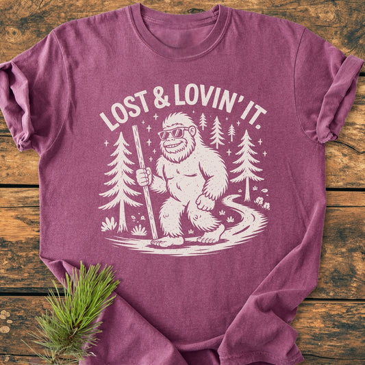 Lost And Lovin' It - Vintage Wash Tee: A purple heathered T-shirt featuring Bigfoot hiking among pine trees, wearing sunglasses, with LOST & LOVIN’ IT text. Laid on wood with a pine sprig beside.