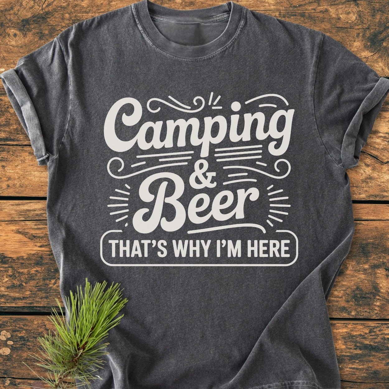Charcoal-gray Camping And Beer - Vintage Wash Tee on wood surface with rolled sleeves, features white text. Pine sprig enhances camping theme. Relaxed fit, soft-washed, made of 100% ring-spun cotton.