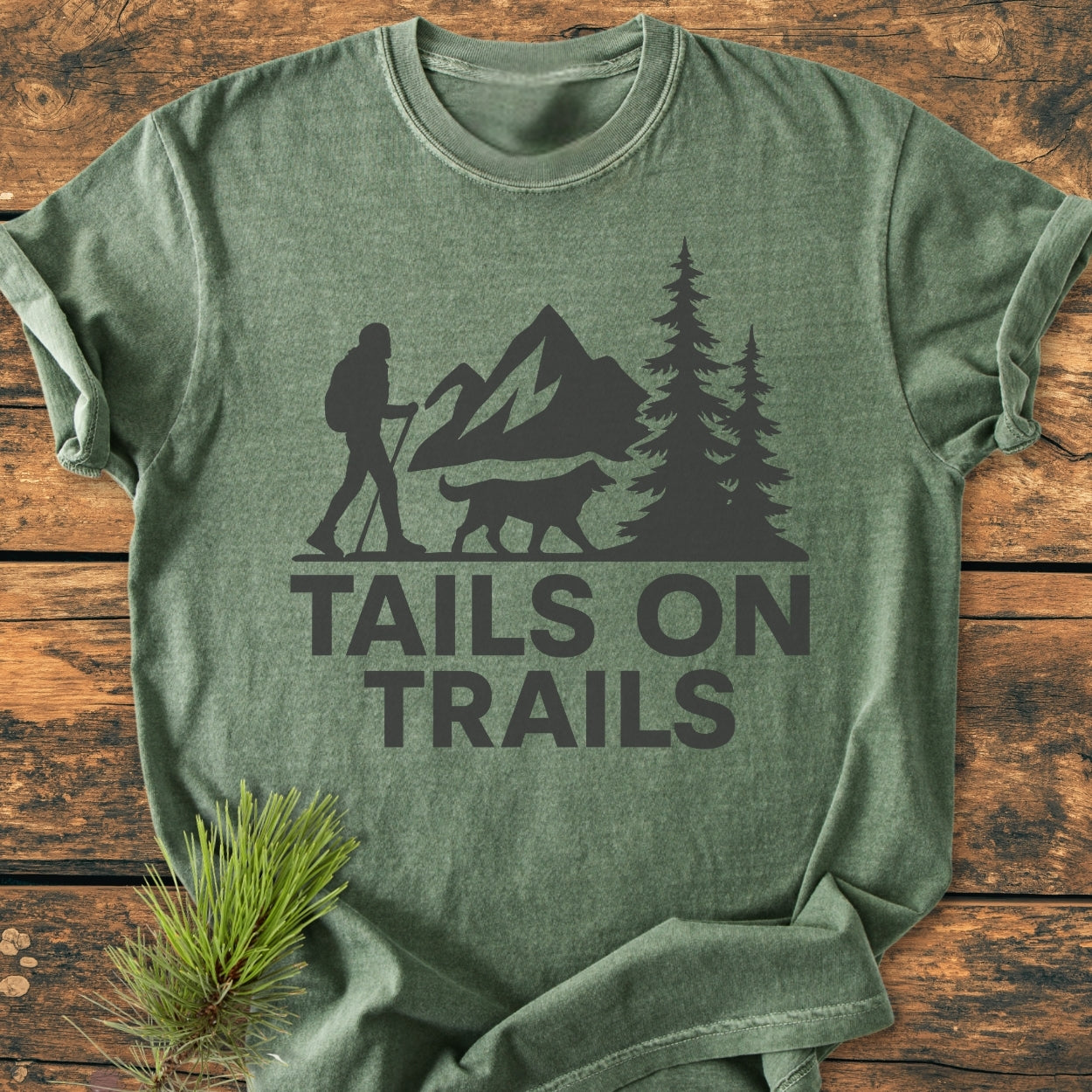 Tails On Trails - Vintage Wash Tee featuring a hiker and dog silhouette with mountains, printed on a green T-shirt, laid on wood. Beside it, a small pine sprig enhances the outdoor theme.