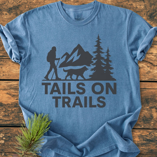 Tails On Trails - Vintage Wash Tee features a hiker and dog silhouette against mountains. This heather-blue tee, on a wood surface, emphasizes comfort with 100% ring-spun cotton and durable stitching.